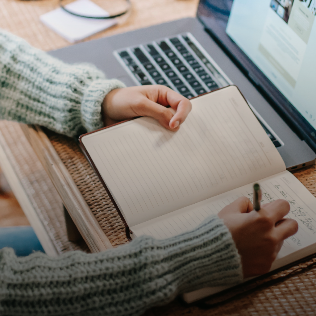 Una persona escribe en un cuaderno abierto mientras trabaja en su computadora portátil, que está encendida y muestra una página web.