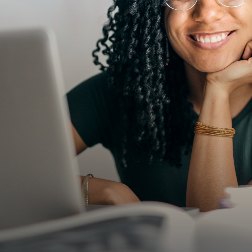 Una mujer sonriente, con el mentón apoyado en su mano, trabaja en su computadora. 