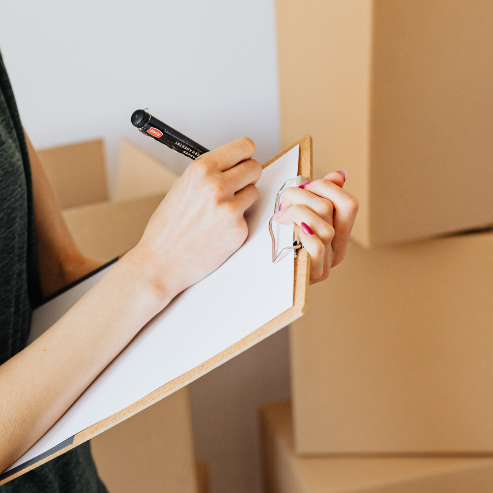 ersona sosteniendo un portapapeles y escribiendo con un marcador, rodeado de cajas de cartón, simbolizando la preparación y organización de inventario o logística en un entorno de almacén o exportación.