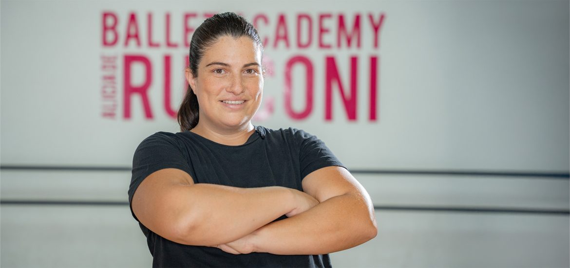Mujer con cabello recogido y camiseta negra, de brazos cruzados, sonríe con confianza en un estudio de ballet. En el fondo, un letrero parcialmente visible con las palabras "Ballet Academy" y "Rugoni" en letras rosadas sobre una pared blanca.