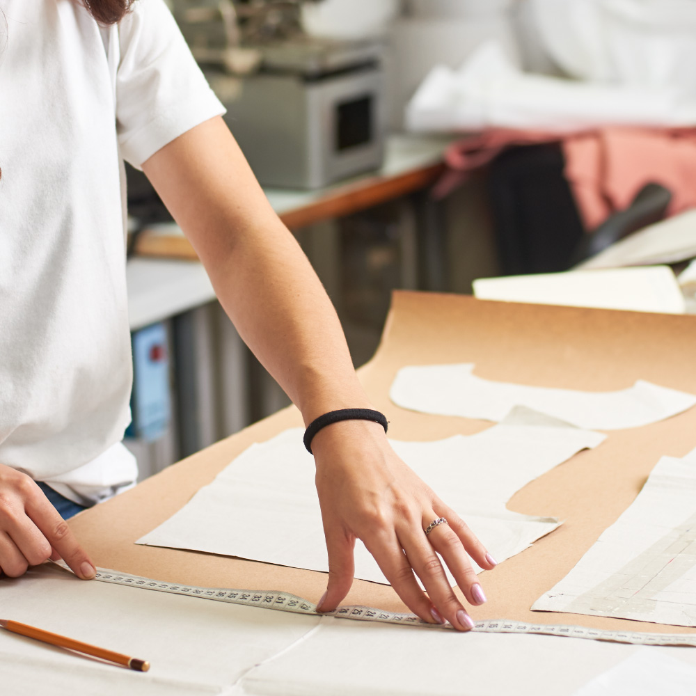 Manos de una persona trabajando en un taller de costura, midiendo y cortando tela sobre una mesa.