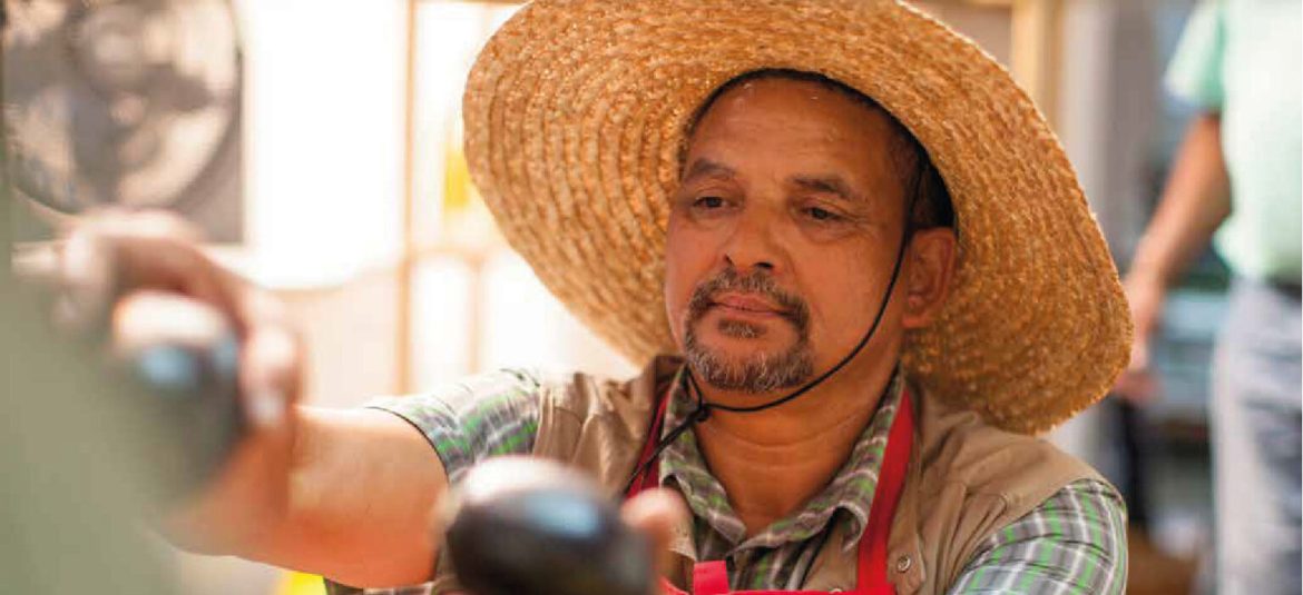 Hombre de mediana edad, con barba y bigote, viste una camisa a cuadros y un sombrero de paja, trabajando en lo que parece ser un mercado o puesto de venta. Está seleccionando productos con cuidado, mostrando una expresión concentrada y dedicada.