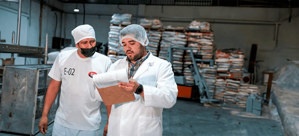 Dos trabajadores, posiblemente en un entorno de fábrica o planta de producción, revisando documentos. Ambos llevan equipo de protección, incluyendo redes para el cabello, batas y uno de ellos una mascarilla. En el fondo se ven varios sacos apilados, lo que indica que se encuentran en un ambiente de almacenamiento o producción de bienes.