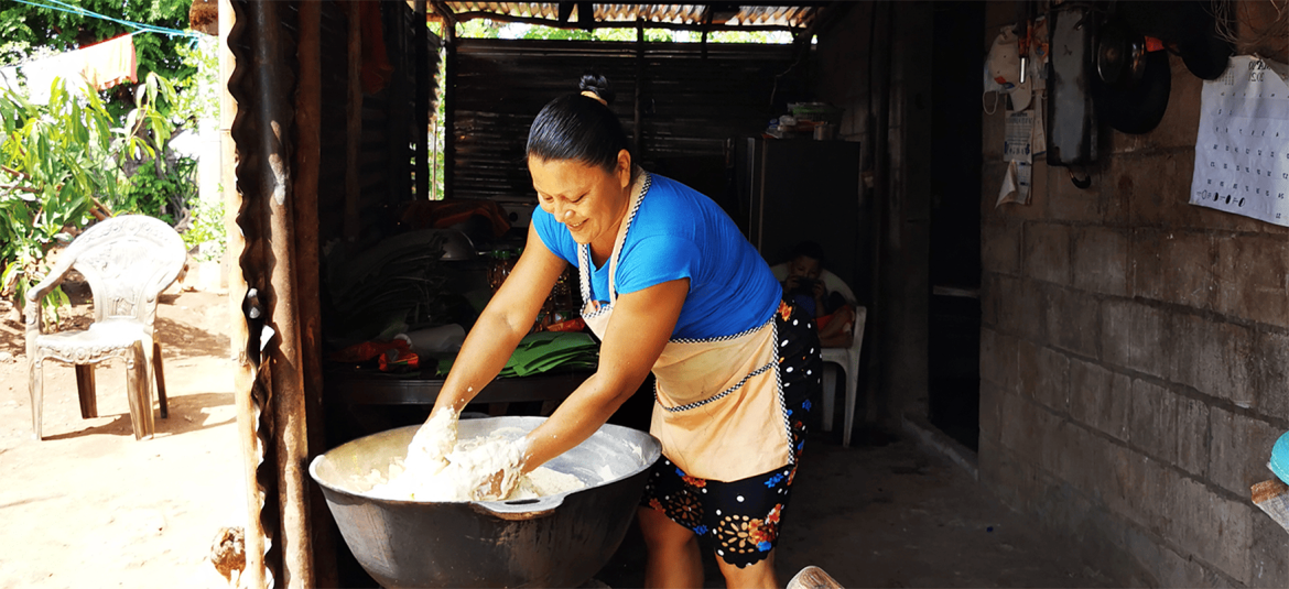 La imagen que has subido muestra a una mujer amasando en un recipiente grande, en lo que parece ser un entorno rural. La escena transmite un ambiente de trabajo artesanal, con énfasis en la labor manual y tradicional. La luz natural entra en la vivienda, iluminando el espacio de trabajo.