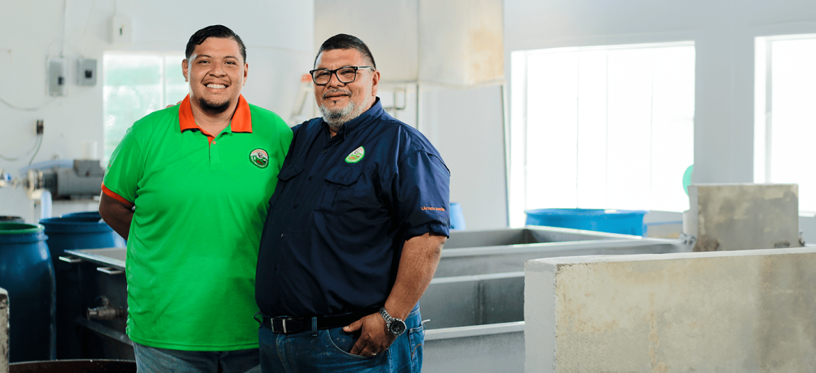 La imagen muestra a dos hombres empresarios, en un entorno industrial o de producción. Ambos están sonriendo y posando juntos, lo que refleja un ambiente de colaboración y éxito. El lugar parece ser una instalación limpia y bien iluminada, con grandes contenedores y maquinaria en el fondo, lo que sugiere un ambiente de trabajo o una fábrica.
