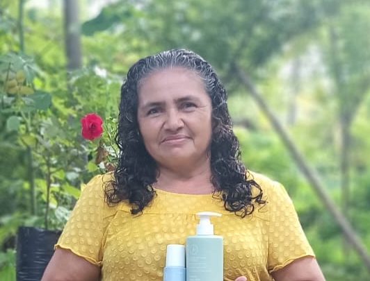 Una mujer de cabello rizado y canoso, vestida con una blusa amarilla, sonríe mientras sostiene productos cosméticos. 