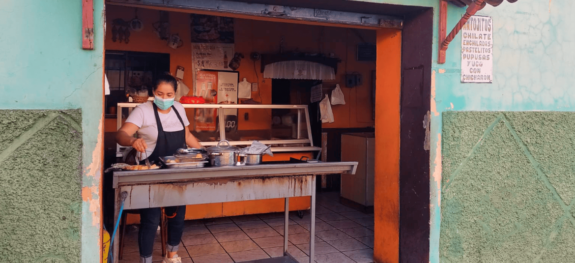 Una mujer preparando comida en un puesto local, probablemente una pequeña fonda o venta de comida típica. La mujer, usando mascarilla y delantal, está frente a una mesa con varias ollas y utensilios de cocina. El local tiene un aspecto sencillo y tradicional, con una pequeña ventana que da al exterior, donde se encuentran los clientes. En la pared hay un letrero que enumera los productos disponibles, como pupusas, pasteles y enchiladas.