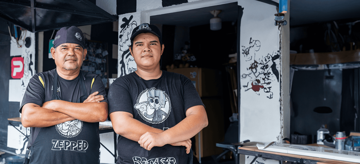 Dos hombres de mediana edad, uno mayor y otro más joven, posan con los brazos cruzados frente a un establecimiento decorado con arte urbano en blanco y negro. Ambos llevan camisetas con logotipos relacionados con su negocio. El hombre mayor luce un delantal negro con la palabra 'ZEPPED' en el frente. Ambos miran con confianza hacia la cámara, simbolizando el esfuerzo y la dedicación empresarial.