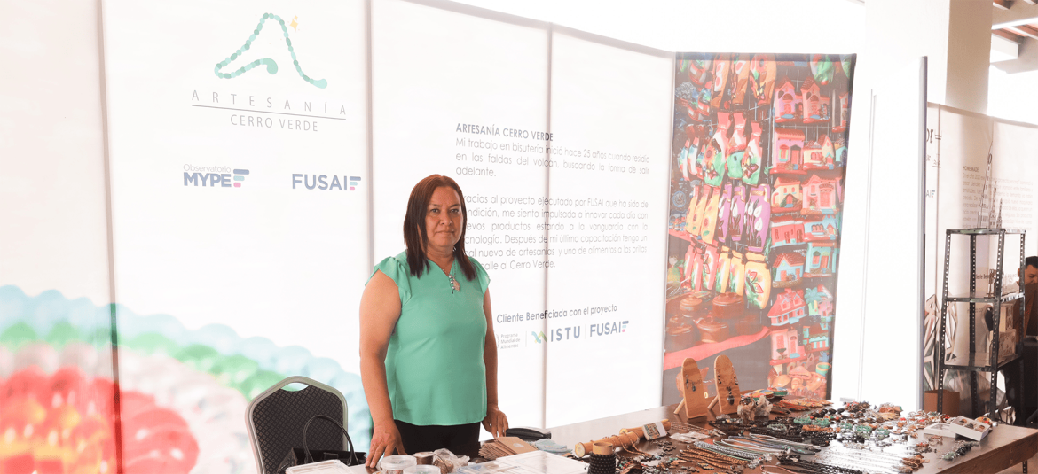 Una mujer se encuentra de pie junto a una mesa exhibiendo diversos productos de bisutería y artesanías. Lleva una blusa verde y su expresión es de orgullo y determinación. Detrás de ella, hay un panel de fondo que describe su emprendimiento, "Artesanía Cerro Verde", y muestra los logotipos de FUSAI y el Observatorio MYPE. También se puede ver una estantería con productos artesanales coloridos, que resaltan la dedicación y creatividad en su trabajo. La imagen ilustra la labor de emprendedores locales apoyados por programas de desarrollo, mostrando su esfuerzo por innovar y mantener sus productos a la vanguardia en un contexto turístico y artesanal.