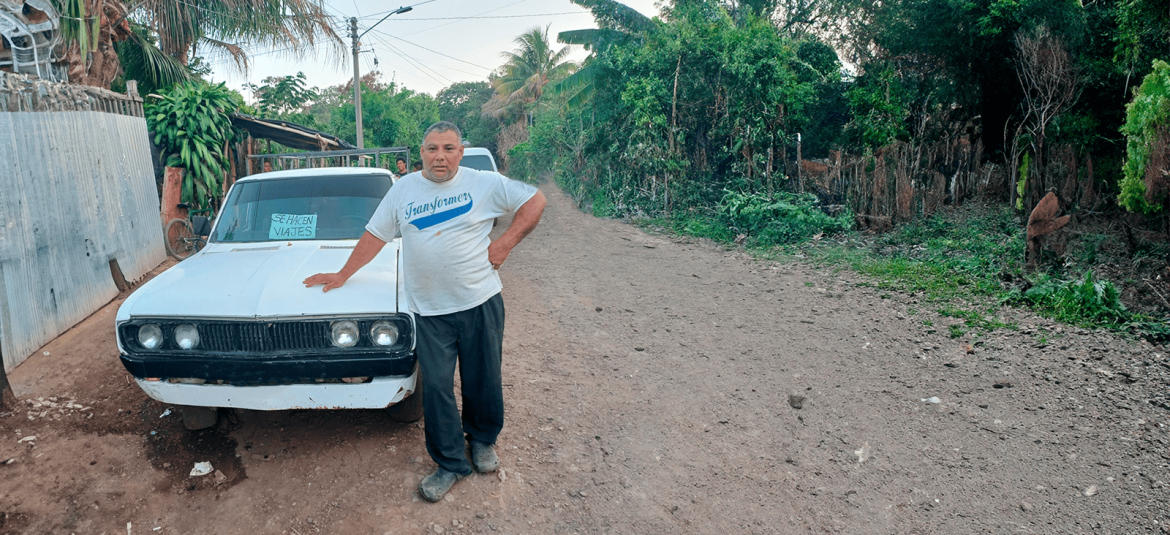 La imagen muestra a un hombre de pie junto a un automóvil antiguo blanco en una calle de tierra rodeada de vegetación densa. El hombre, vestido con una camiseta que dice "Transformers" y pantalones oscuros, descansa una mano sobre el capó del automóvil, en cuyo parabrisas delantero hay un letrero que dice "Se hacen viajes", lo que indica que probablemente ofrece servicios de transporte o taxi informal. El entorno sugiere una zona rural, con una mezcla de vegetación y cercas sencillas, creando una atmósfera tranquila y cotidiana.