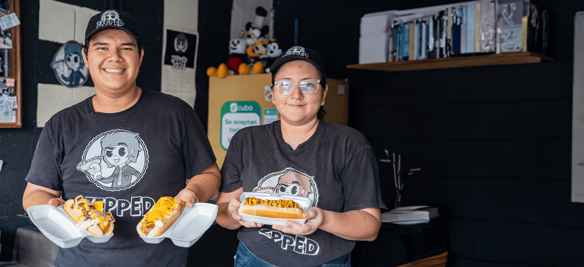 La imagen muestra a dos personas jóvenes, un hombre y una mujer, quienes sonríen mientras sostienen platos de comida, específicamente hot dogs cubiertos con salsas. Ambos llevan camisetas y gorras negras con el logo de su negocio, que parece llamarse "Zapped". Están en lo que parece ser el interior de su local, con una decoración informal y algunos elementos visibles en el fondo, como una estantería con libros y muñecos decorativos. La imagen transmite un ambiente acogedor y amigable, resaltando el entusiasmo de los jóvenes emprendedores por su negocio de comida.