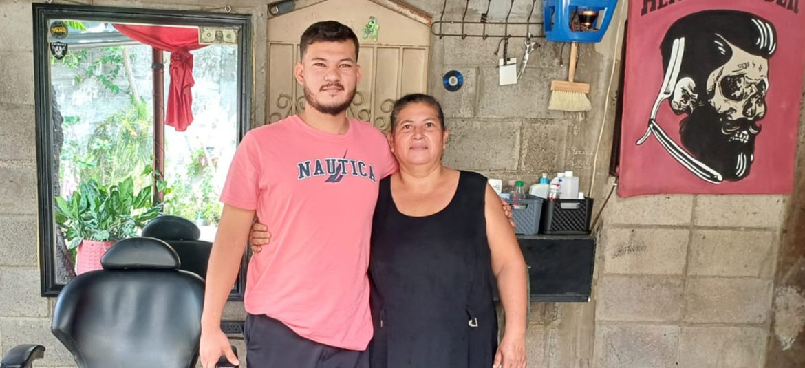 La imagen muestra a un joven y a una mujer, posiblemente madre e hijo, en el interior de lo que parece ser una barbería o salón de peluquería. El joven, con una camiseta rosada que dice "Nautica," tiene un brazo alrededor de la mujer, quien viste un vestido negro y muestra una expresión amigable. En el fondo se puede ver un espejo grande, una silla de barbero y productos de cuidado personal organizados en estantes. La decoración incluye una cortina roja con el diseño de una calavera estilizada y una navaja de barbero, lo que refuerza el ambiente de barbería.