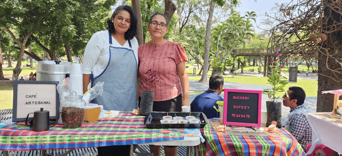 La imagen muestra a dos mujeres en un puesto de ventas al aire libre, rodeadas de árboles y en un entorno de parque. La mujer a la izquierda lleva un delantal azul, indicando que está involucrada en la preparación o venta de productos, mientras que la mujer a la derecha lleva una blusa rosada y parece estar apoyando la exhibición. En la mesa, cubierta con un mantel colorido, se ofrecen muestras de comida y café, con letreros que dicen "Café Artesanal" y "Bakery Sofkey" junto con un número de contacto. La decoración incluye un frasco con granos de café y una pequeña planta, lo que da un toque artesanal y natural al ambiente.