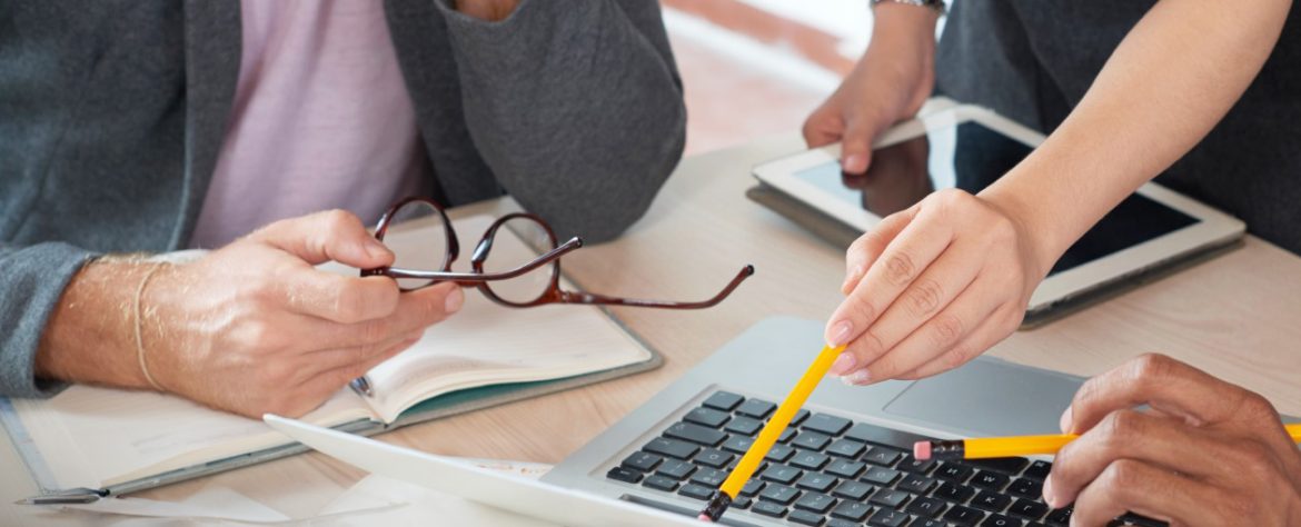 La imagen muestra un grupo de personas trabajando juntas alrededor de una computadora portátil. Una persona sostiene un par de gafas, mientras otra señala la pantalla de la computadora con un lápiz, indicando un enfoque colaborativo en la tarea. En la mesa también hay una libreta abierta, una tableta y otros instrumentos de trabajo, lo que sugiere un entorno de discusión o planificación, probablemente relacionado con la creación o análisis de estrategias. El ambiente es profesional y de trabajo en equipo.