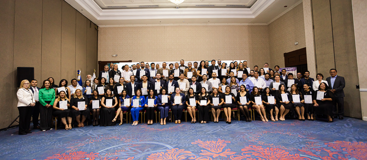 Grupo numeroso de personas posando con certificados en mano durante una ceremonia de graduación empresarial organizada por FUSAI. Los participantes, en su mayoría vestidos de manera formal, están divididos en varias filas dentro de un salón de eventos. Al fondo se pueden ver banderas y pancartas, destacando el éxito de los programas de activación empresarial