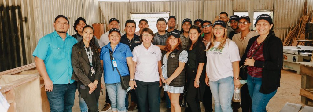 Grupo de personas posando para una foto en un taller, todos vestidos de manera casual. Algunos llevan gorras con el logotipo de FUSAI, lo que sugiere que están participando en una actividad organizada por la institución. El ambiente parece ser el interior de un espacio de trabajo o capacitación