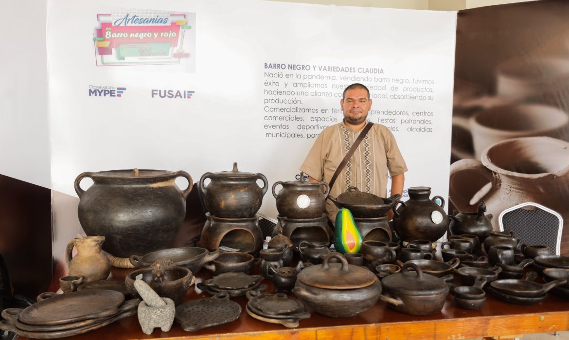 La imagen muestra a un hombre de pie detrás de una mesa llena de artesanías de barro negro. En el fondo, hay un cartel que dice "Artesanías Barro Negro y Rojo, Variedades Claudia" con los logotipos de FUSAI y el Observatorio MYPE. El texto también menciona que el emprendimiento nació en la pandemia, con éxito en la venta de barro negro, y se expandió mediante la creación de una alianza con la comunidad local para absorber su producción. En la mesa, se pueden ver varios artículos de barro como ollas, platos, tazones y otros utensilios artesanales. El ambiente refleja una exhibición artesanal, probablemente en una feria