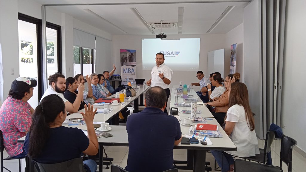 Grupo de empresarios participando en una sesión de capacitación organizada por FUSAI. En la imagen, un facilitador se encuentra de pie frente a los participantes, quienes levantan la mano para intervenir, en un entorno colaborativo. En la pantalla se visualiza el logo de FUSAI y MBA Micro. Alrededor de la mesa hay cuadernos y tazas de café, lo que resalta el ambiente participativo y profesional de la reunión.