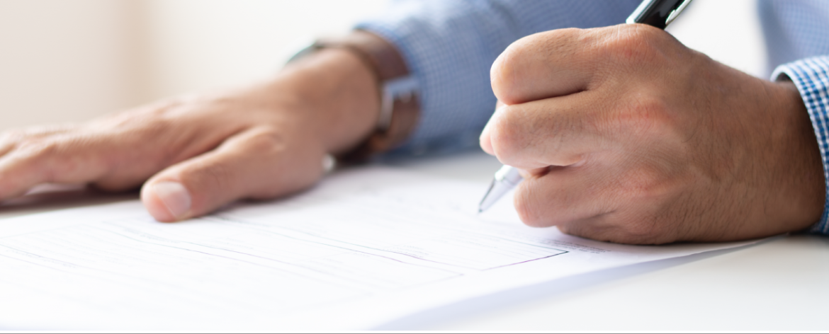 Primer plano de una mano masculina llenando un formulario o documento oficial con un bolígrafo. La persona lleva una camisa azul de manga larga y un reloj, lo que sugiere un entorno profesional o de negocios.