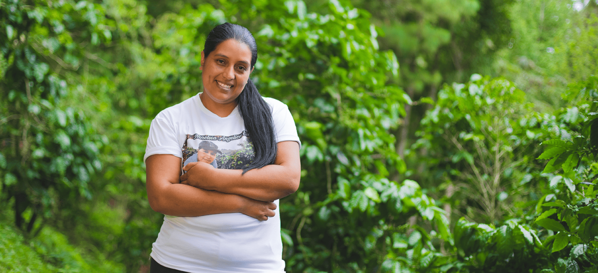 La imagen muestra a una mujer de pie al aire libre, rodeada de abundante vegetación, lo que sugiere un entorno natural o rural. Ella viste una camiseta blanca con una fotografía impresa en el pecho y sonríe de manera amable, con los brazos cruzados de manera relajada. Su expresión refleja confianza y serenidad, en un contexto lleno de plantas verdes que resaltan el ambiente fresco y natural a su alrededor.