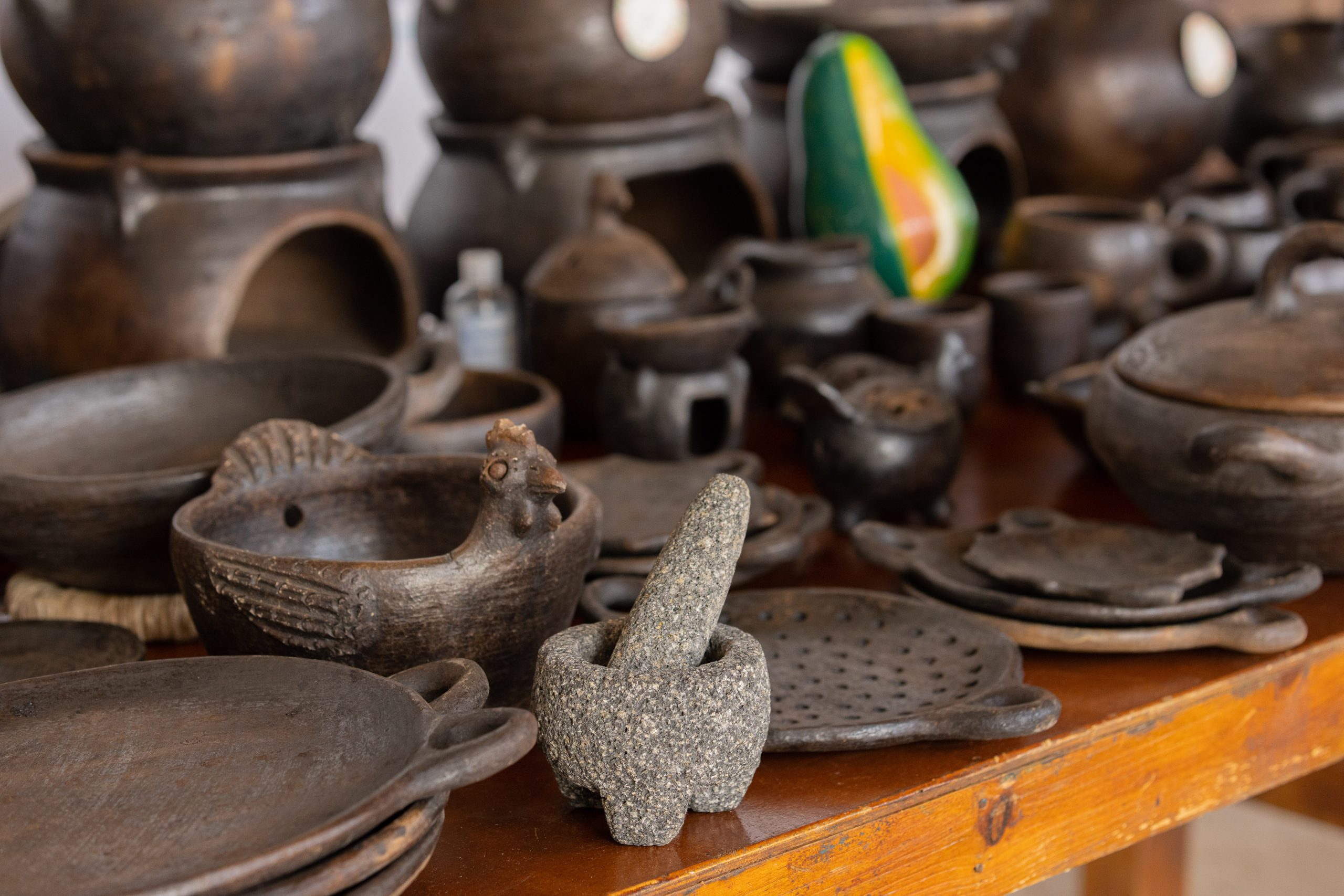 Una variedad de artesanías de cerámica tradicionales de El Salvador dispuestas sobre una mesa. Entre los objetos, destacan platos, ollas y utensilios, como un mortero con su mano de piedra, además de una figura en forma de gallina hecha de barro. Al fondo se observan más recipientes de cerámica y un adorno en forma de aguacate