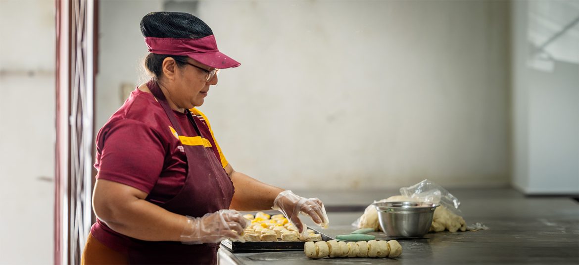 Mujer con uniforme de trabajo y gorra en una cocina, preparando productos horneados en una bandeja.