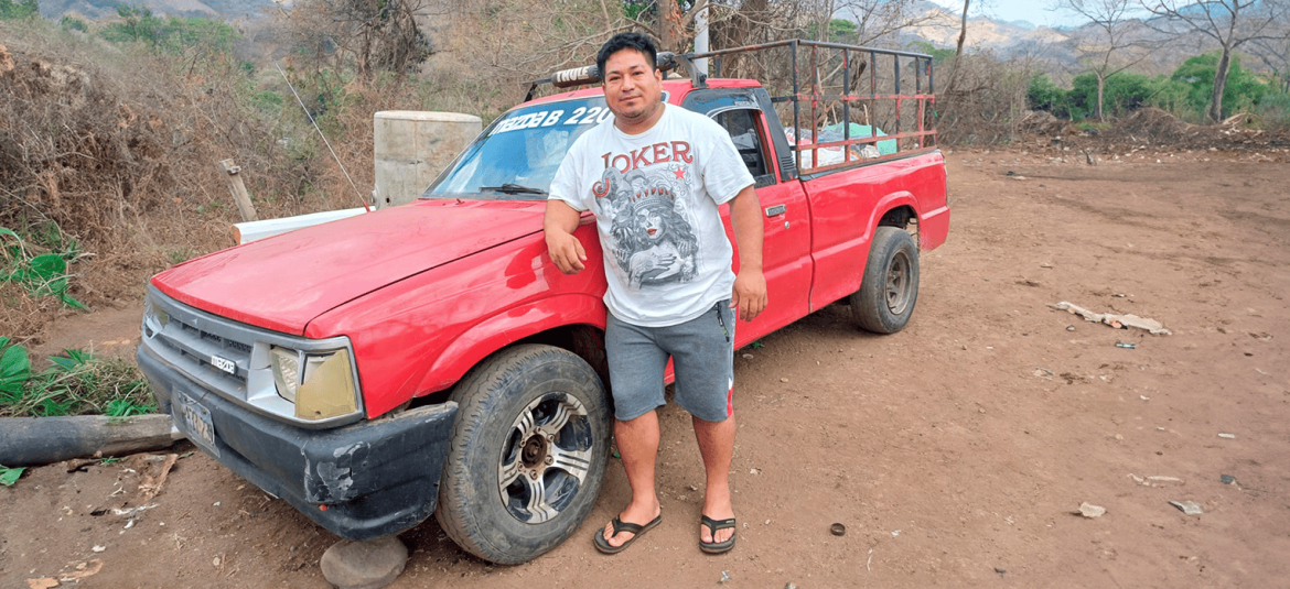La imagen muestra a un hombre apoyado en una camioneta roja, de aspecto antiguo y con señales de uso, en un entorno rural. El hombre viste una camiseta blanca con un diseño gráfico y pantalones cortos grises, junto con sandalias. La camioneta tiene una estructura en la parte trasera, lo que sugiere que podría utilizarse para el transporte de carga o materiales en la zona. El paisaje alrededor es árido, con vegetación seca y algunas montañas al fondo, reflejando un ambiente de trabajo en áreas rurales o semirrurales.