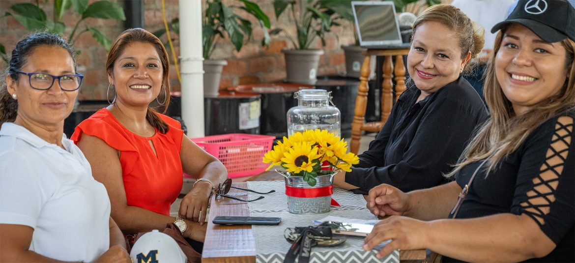 La imagen muestra a cuatro mujeres sentadas en una mesa, compartiendo un momento de conversación y sonrisas en un ambiente acogedor. En el centro de la mesa hay un arreglo de girasoles en una pequeña maceta decorativa, que añade color y calidez al ambiente. Las mujeres están vestidas de manera casual y se ven relajadas, lo que sugiere un entorno amistoso y ameno. Al fondo, se observan plantas y una computadora portátil, lo que podría indicar un espacio de trabajo o reunión informal.