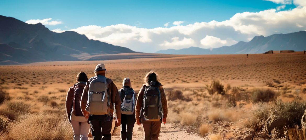 La imagen muestra a un grupo de cuatro personas caminando por un paisaje desértico hacia un horizonte amplio y montañoso. Están equipados con mochilas, ropa cómoda y se dirigen hacia las montañas en la distancia. La escena refleja una atmósfera de exploración y aventura, resaltando la inmensidad del terreno árido que los rodea, con arbustos bajos y un cielo despejado, salvo por algunas nubes. La imagen transmite una sensación de travesía, trabajo en equipo y conexión con la naturaleza en un entorno vasto y desolado.