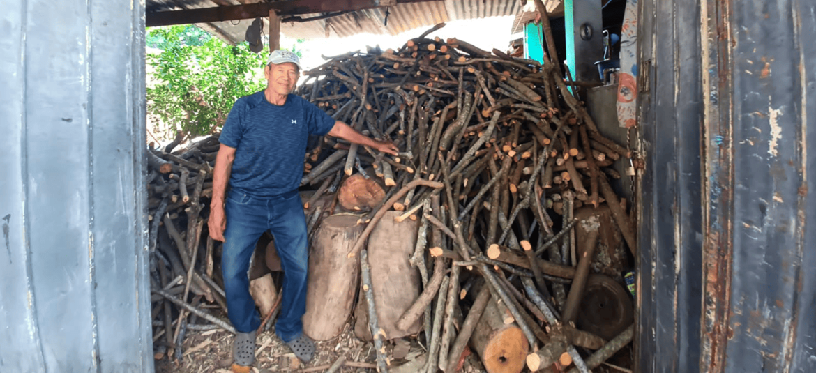 la-dinámica-empresarial-en-los-segmentos-de-la-micro-y-pequeña-empresa La imagen muestra a un hombre mayor, de pie junto a una gran pila de leña apilada en el interior de una estructura rústica. Él viste una camiseta azul y pantalones de mezclilla, y lleva una gorra. Su mano descansa sobre la leña, indicando su trabajo o relación con la actividad de recolección y almacenamiento de leña. La pila de troncos y ramas es considerable y está organizada de manera que ocupa gran parte del espacio, sugiriendo que es un recurso esencial para la actividad que se realiza en el lugar.