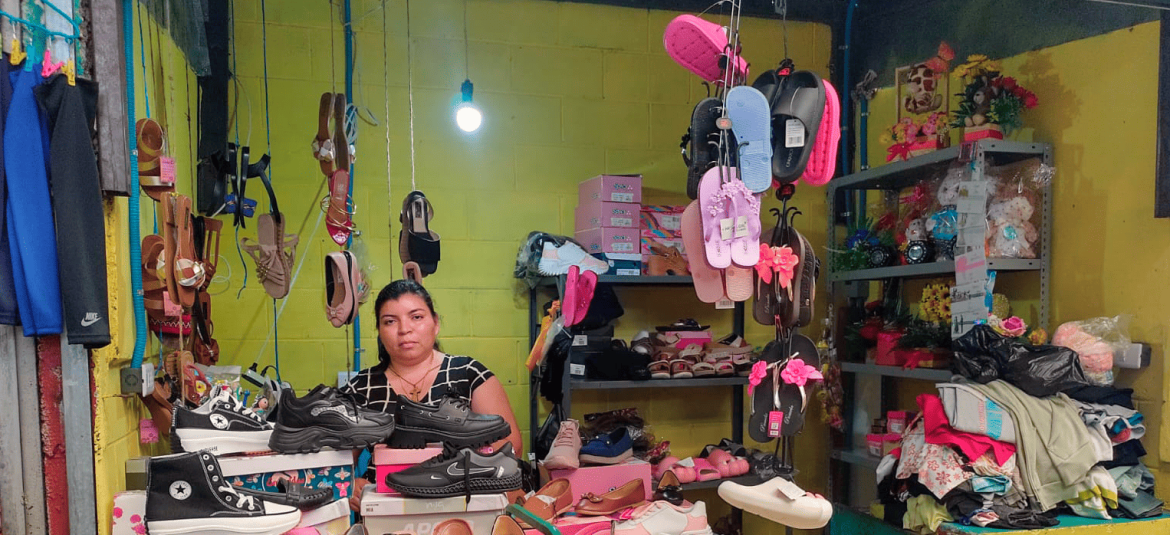 micro-y-pequeños-empresarios-no-formales-recuperan-confianza-en-la-economía La imagen muestra a una mujer en un pequeño negocio de venta de calzado y otros artículos, en lo que parece ser un mercado o tienda informal. Está rodeado de varios productos, incluidos zapatos deportivos, sandalias y calzado de varios estilos, que están organizados en estantes y colgados alrededor del puesto. Las paredes del espacio son de color amarillo brillante, y el ambiente transmite una atmósfera de sencillez y esfuerzo por mostrar los productos de la mejor manera posible. La mujer tiene una expresión seria, reflejando quizás concentración o dedicación en su trabajo.
