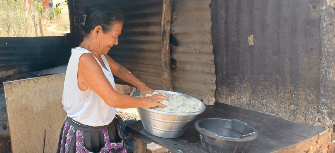 La imagen muestra a una mujer trabajando al aire libre, posiblemente en una actividad culinaria tradicional. Está amasando una gran cantidad de masa en un recipiente metálico, lo que sugiere que está preparando alimentos en un entorno rústico o rural. La mujer viste una blusa blanca sin mangas y una falda con detalles decorativos. A su alrededor se observan paredes de lámina y algunos elementos de su entorno, que indican que el lugar es un espacio sencillo y funcional.