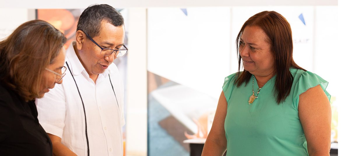 La imagen muestra a tres personas interactuando en lo que parece ser una feria o exposición de productos. Dos mujeres y un hombre observan varios artículos sobre una mesa, posiblemente analizando productos artesanales o alimenticios en pequeños envases. El hombre, que lleva una camisa blanca, está señalando algo en la mesa, mientras que las dos mujeres, una de camisa verde y otra de color oscuro, lo observan con atención. La situación refleja un ambiente de compra o evaluación de productos, probablemente relacionado con emprendimientos locales o microempresas.