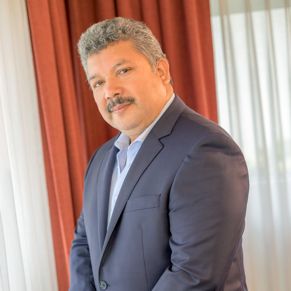 Hombre de mediana edad con traje formal posando frente a una ventana. Lleva una chaqueta oscura y camisa clara, proyectando una imagen profesional y seria. Al fondo, se observan cortinas en tonos rojos que contrastan con la iluminación suave de la escena.