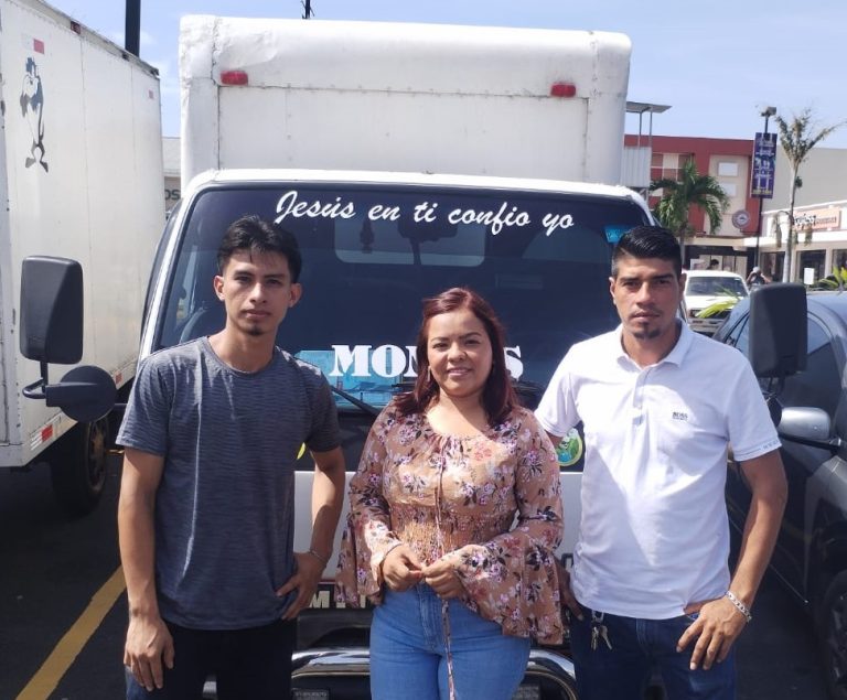 Tres personas posando frente a un camión de carga con un mensaje en el parabrisas que dice "Jesús en ti confío yo". A la izquierda, un hombre joven con camiseta gris. En el centro, una mujer con blusa estampada floral y cabello suelto. A la derecha, un hombre con camisa blanca de pie. Están en un estacionamiento, con algunos edificios y árboles visibles al fondo.