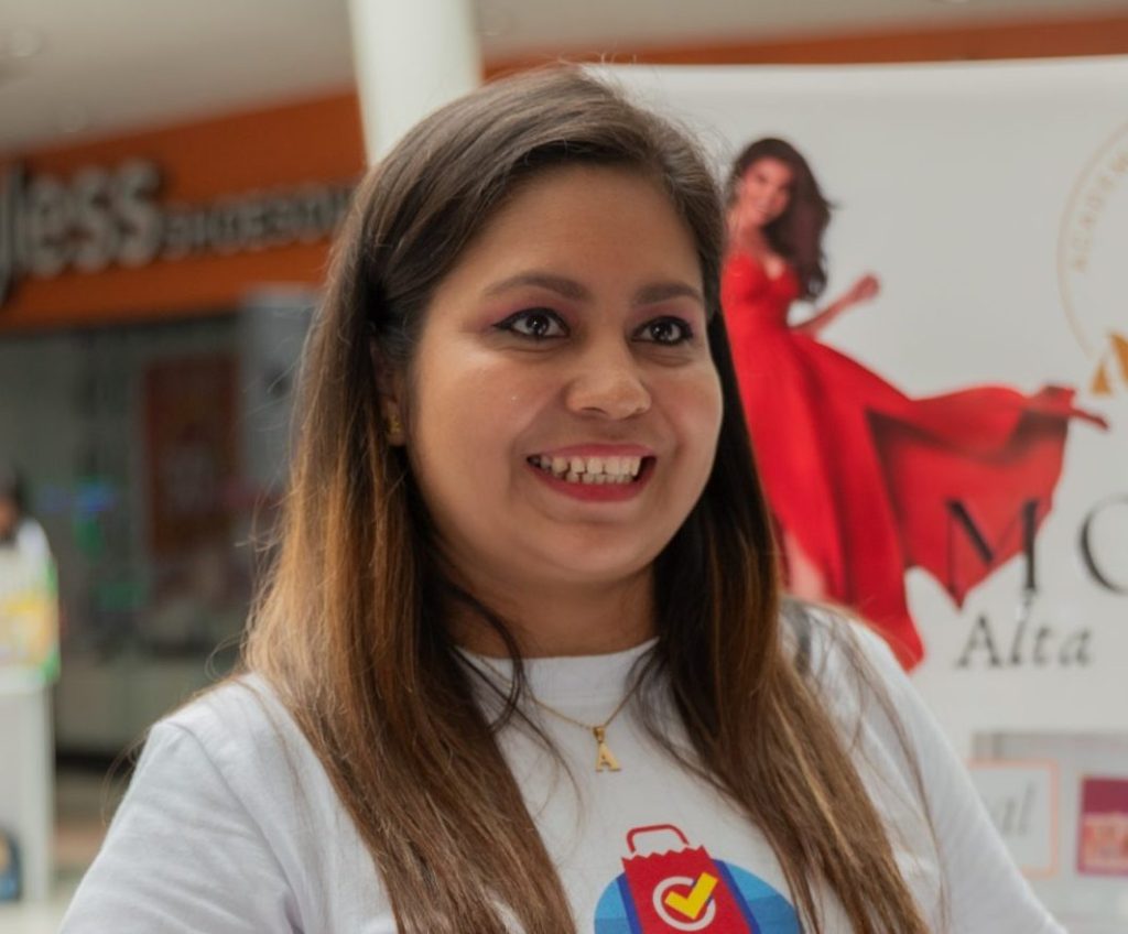 Una mujer sonriente con el cabello largo y lacio está en un evento, vistiendo una camiseta con un diseño colorido en el centro. 