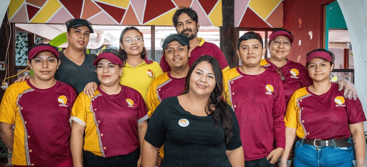 Un grupo de personas posa para la foto, mostrando un ambiente de trabajo en equipo y camaradería. La mayoría de los integrantes visten uniformes en tonos rojo y amarillo con viseras, identificando su pertenencia a una misma empresa o establecimiento, mientras que una mujer en el centro lleva una blusa negra y una credencial. La pared del fondo presenta un diseño colorido que agrega un toque de vitalidad y dinamismo. La imagen transmite un mensaje de unidad, colaboración y orgullo por el equipo, ideal para ilustrar temas de trabajo en equipo, cohesión laboral o identidad de marca.