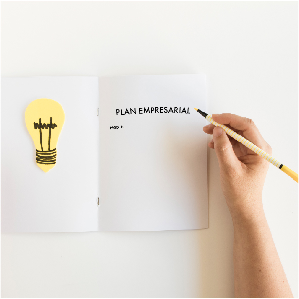 Mano sosteniendo un lápiz amarillo y escribiendo en un cuaderno abierto que dice 'Plan Empresarial - Paso 1'. Al lado izquierdo de la página hay una ilustración de una bombilla amarilla que simboliza una idea.