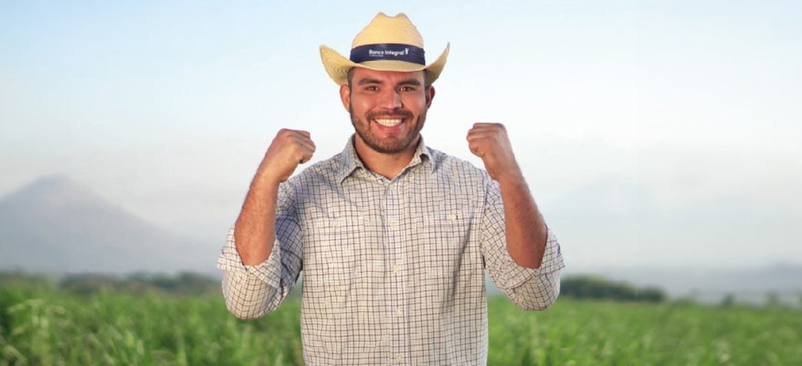 La imagen muestra a un hombre joven, con una actitud positiva y enérgica, levantando ambos puños en señal de entusiasmo y éxito. Lleva una camisa a cuadros y un sombrero con el logo de "Banco Integral". El fondo es un paisaje natural con montañas y vegetación, lo que sugiere un entorno rural. La imagen transmite una sensación de logro y optimismo, probablemente relacionada con el apoyo del Banco Integral hacia los micro y pequeños empresarios en el país, especialmente en áreas rurales o agrícolas.