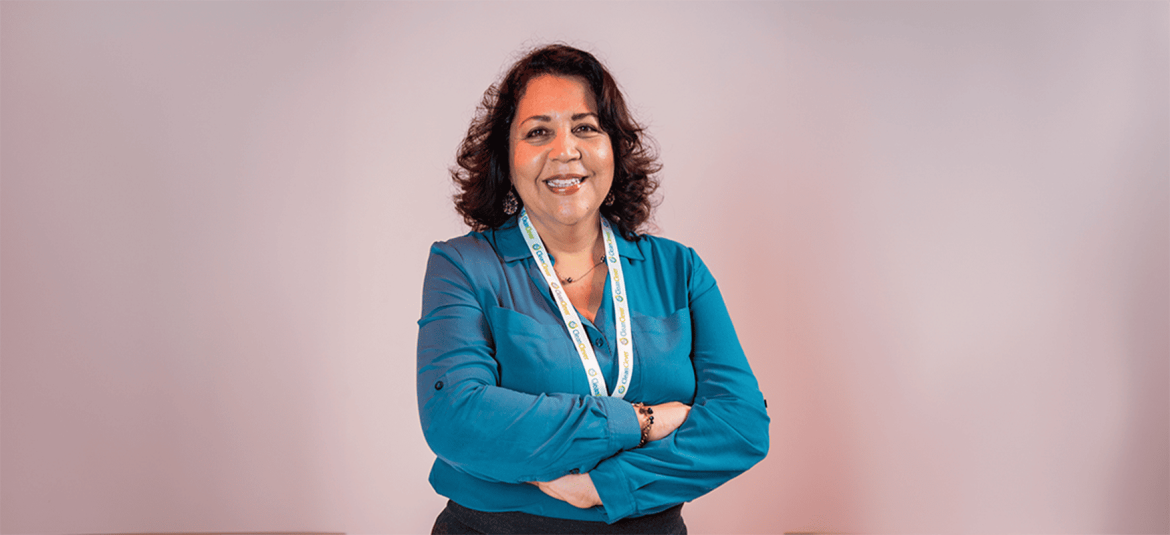 Una mujer de pie, con los brazos cruzados y una sonrisa cálida, viste una camisa azul y porta una credencial en el cuello. El fondo es liso y de color neutro, lo que resalta su figura y expresión. La imagen refleja confianza, profesionalismo y una actitud positiva, ideal para representar temas de liderazgo, asesoría o empoderamiento en el ámbito profesional.