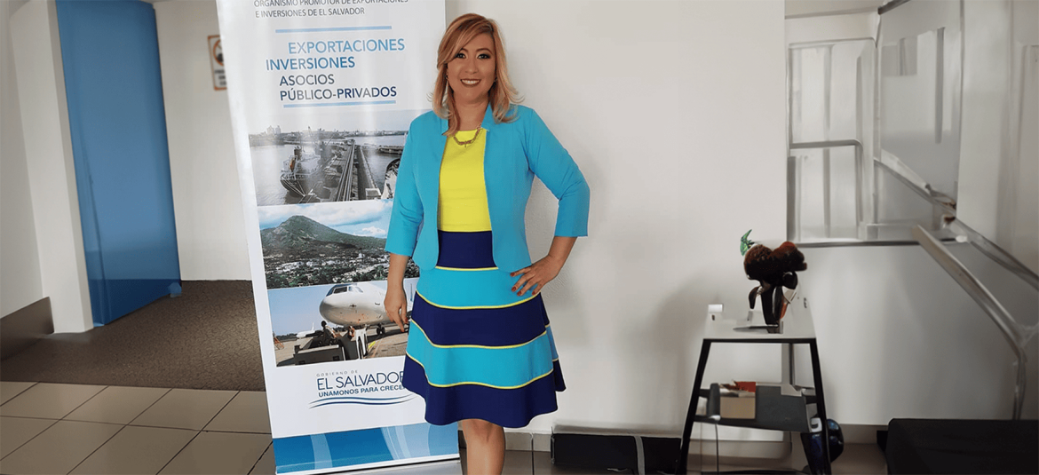 Una mujer sonriente y profesional, vestida con un conjunto colorido en tonos de azul y amarillo, se encuentra de pie junto a una pancarta promocional del Gobierno de El Salvador.
