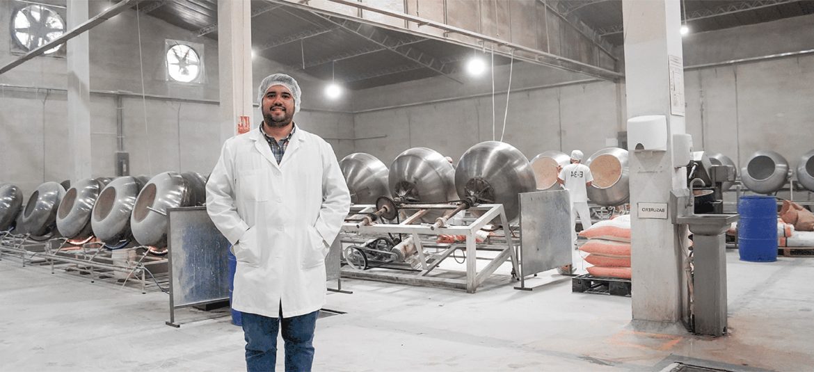 clima-de-confianza-empresarial La imagen muestra a un hombre de pie en una planta de procesamiento industrial, vestido con una bata blanca, redecilla en el cabello y una expresión amigable. Al fondo, se observan varias máquinas de acero inoxidable, que parecen ser tambores de mezcla o procesamiento, alineadas en una fila. Otro trabajador aparece en la parte posterior, también vestido de blanco, supervisando una de las máquinas. La planta es amplia y está bien iluminada, con un ambiente ordenado y limpio que indica un alto estándar de higiene, posiblemente para la producción de alimentos o productos químicos.