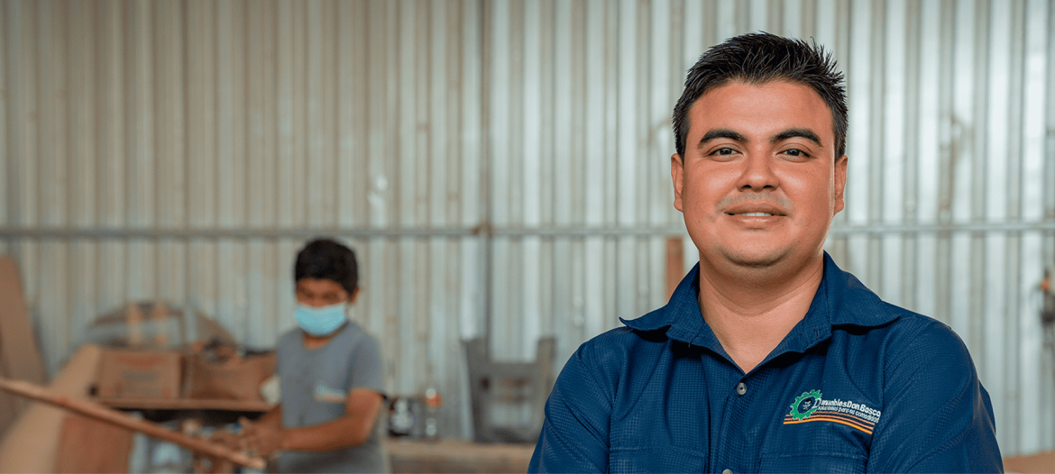 La imagen muestra a un hombre de pie, con una expresión segura y una leve sonrisa, vestido con una camisa azul con logotipo, probablemente representando a una empresa o iniciativa. En el fondo, se observa a otra persona trabajando, posiblemente en tareas de manufactura o producción, usando mascarilla. El entorno parece ser un taller, resaltando el trabajo en equipo y el ambiente industrial. La imagen refleja profesionalismo y dedicación en el ámbito laboral, además de una atmósfera de colaboración en un espacio de trabajo práctico y funcional.