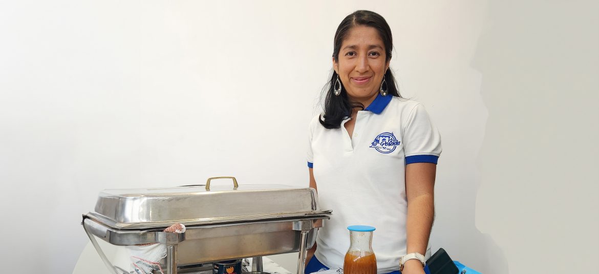 clima-de-confianza-empresarial-segundo-trimestre-2023 Mujer empresaria con cabello oscuro, vestida con una camiseta blanca con el logotipo de 'La Posada' en el pecho, de pie junto a una bandeja de comida metálica cubierta. Frente a ella hay una botella de vidrio con salsa. La imagen sugiere que está relacionada con un negocio de servicios alimentarios
