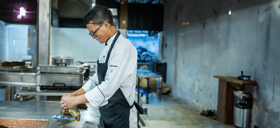 La imagen muestra a un chef concentrado en su trabajo, vistiendo uniforme blanco y un delantal negro, mientras envuelve cuidadosamente un producto en papel dorado sobre una superficie de acero inoxidable. El entorno es una cocina industrial, con equipos de cocina al fondo y una iluminación suave que resalta la dedicación del chef en su tarea. La expresión tranquila y la postura atenta sugieren profesionalismo y precisión en su trabajo.