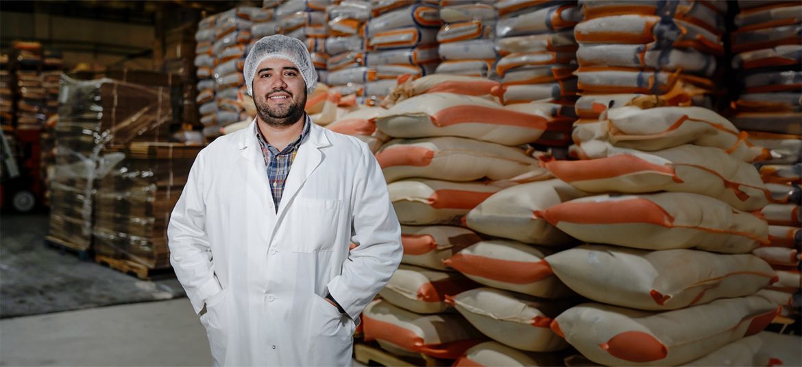 Un hombre con una bata de laboratorio y una malla en el cabello sonríe frente a una pila de sacos apilados en un almacén. La imagen transmite profesionalismo y dedicación en un ambiente industrial, destacando la importancia del control de calidad y el trabajo en equipo en el sector de almacenamiento o producción.