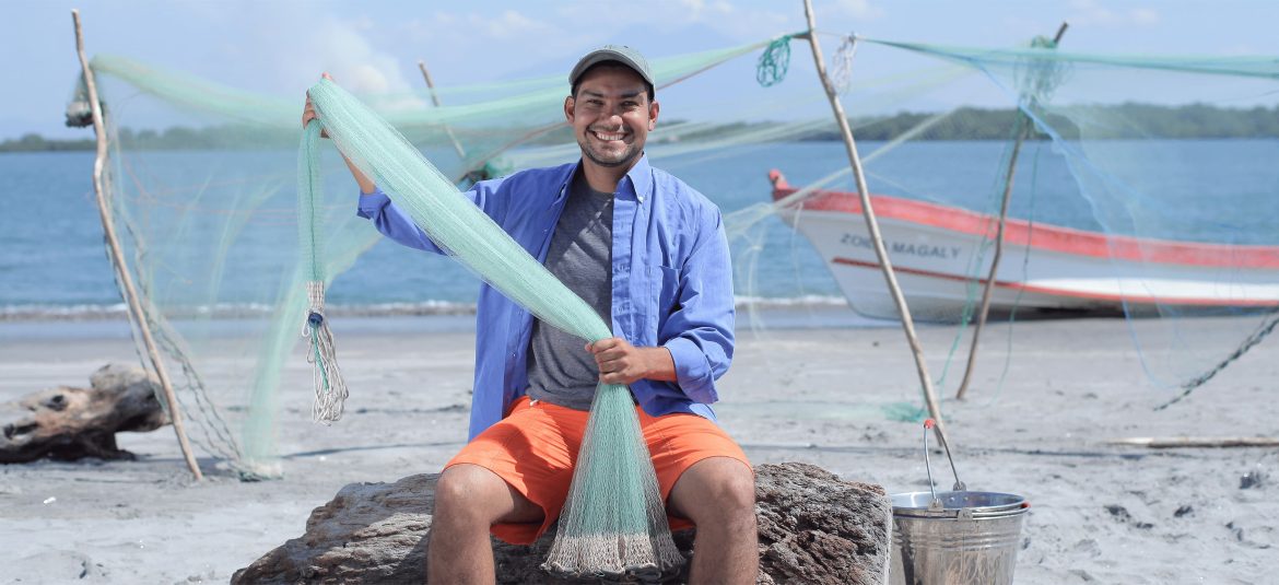 La imagen muestra a un pescador sonriente sentado en la playa mientras sostiene una red de pesca. Lleva puesta una gorra, una camisa azul y pantalones cortos naranjas, lo que le da un aspecto relajado y cómodo para su entorno. Detrás de él, se puede ver el océano y una embarcación con el nombre "Zona Magaly", así como estructuras improvisadas para secar redes. La escena transmite una conexión directa con el mar y refleja la actividad de pesca artesanal en un ambiente costero y natural.
