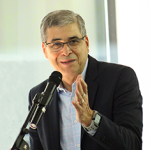 Hombre de mediana edad con cabello canoso, usando gafas y un traje oscuro. Habla frente a un micrófono, haciendo un gesto con una mano, como si estuviera dando una presentación o discurso