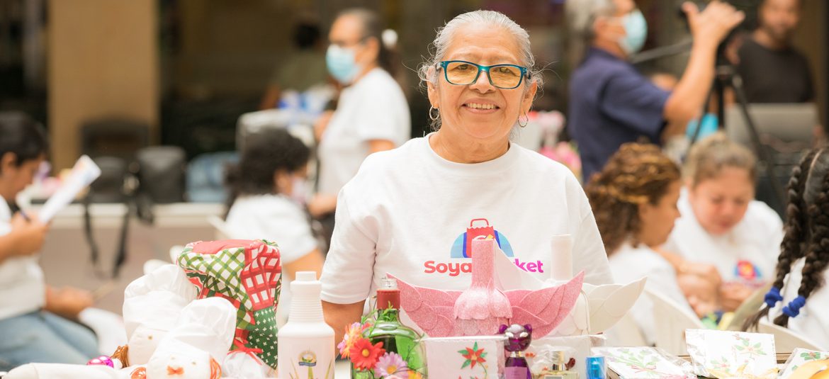 La imagen muestra a una mujer sonriente con cabello canoso y gafas, vestida con una camiseta blanca que tiene el logotipo de "Soyapango Market". Frente a ella hay una variedad de productos artesanales, coloridos y bien organizados sobre una mesa. Detrás, se observan otras personas participando en el evento, algunas usando mascarillas. La escena transmite un ambiente de mercado o feria, enfocada en productos locales y la microempresa, resaltando el esfuerzo y dedicación de las emprendedoras en su labor artesanal.