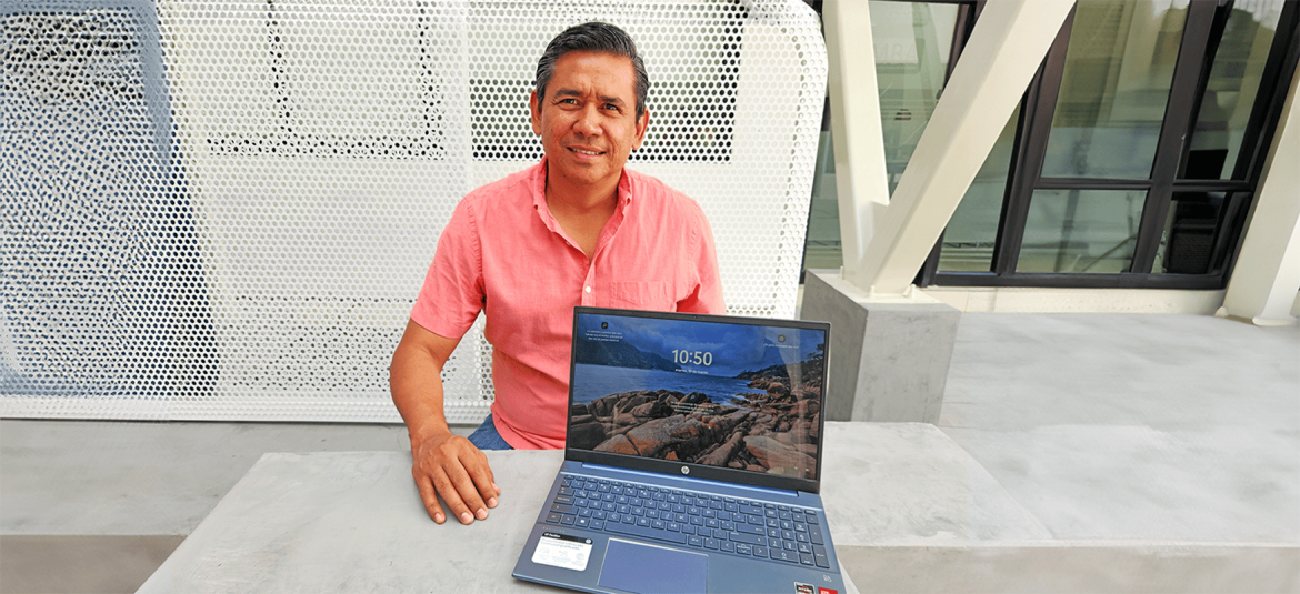 Un hombre con camisa rosada, sentado al aire libre, sonríe mientras tiene una computadora portátil abierta frente a él sobre una mesa.